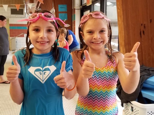 Two girls in swimsuits and swim goggles smiling and giving thumbs up.